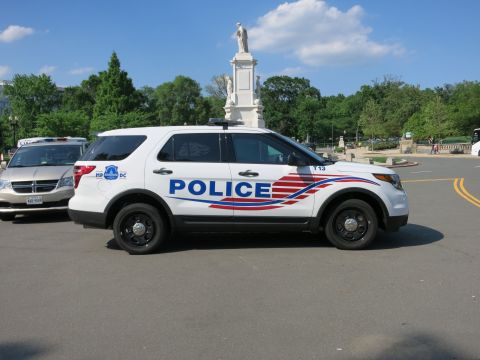 1991 Ford LTD Crown Victoria - white Ford Explorer marked POLICE with red and blue swoosh striping parked under blue sky beside a tall statue.