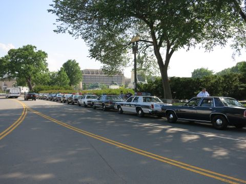 1991 Ford LTD Crown Victoria - row of vintage police sedans and a black Ford LTD-style sedan parked single file along a curb under shade trees.
