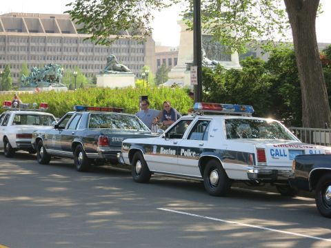 1991 Ford LTD Crown Victoria - Vintage Ford LTD Crown Victoria police cruisers, including Boston Police and State units, parked in a row by a hedge and statues.