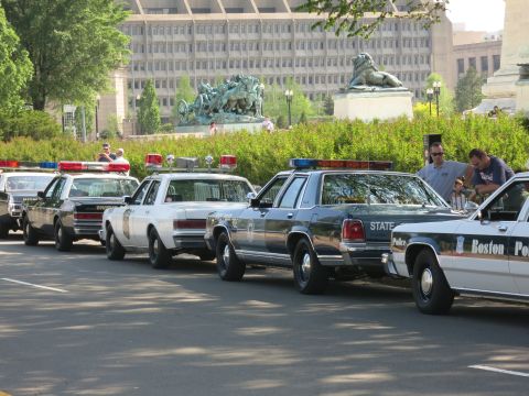 1991 Ford LTD Crown Victoria - line of older State Police and Boston Police Ford LTD-style cruisers parked along a road beneath bronze monuments.