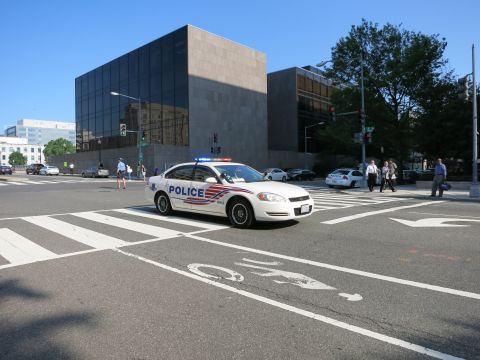 1991 Ford LTD Crown Victoria - white Chevrolet Impala police sedan marked POLICE crossing a crosswalk at an intersection near a dark modern building.