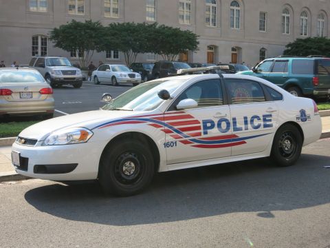 1991 Ford LTD Crown Victoria - white Chevrolet Impala marked POLICE 1601 with patriotic swoosh graphics parked beside a stone building and lot.