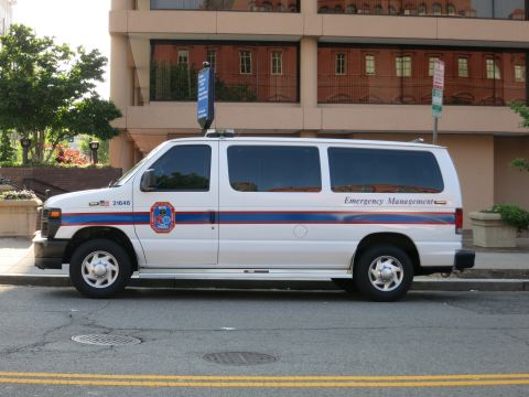1991 Ford LTD Crown Victoria - white Ford E-Series van marked Emergency Management with red and blue striping is parked along a curb beside a brick building.