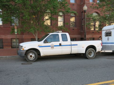 1991 Ford LTD Crown Victoria - white Ford dually Police pickup truck connected to a horse trailer is parked along a street in front of a brick building.