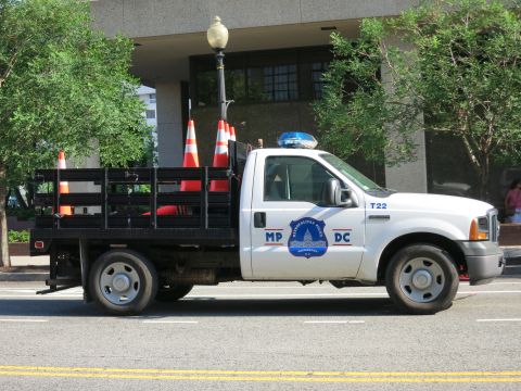 1991 Ford LTD Crown Victoria - white Ford flatbed stake-bed truck marked MPDC, loaded with orange traffic cones, parked on a city street.