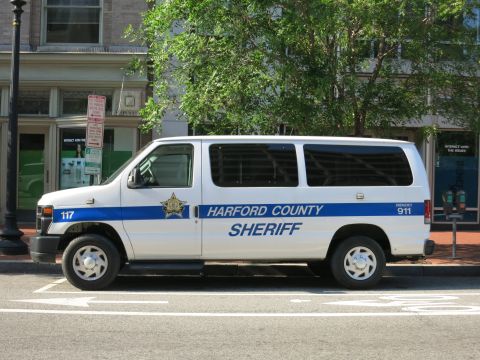 1991 Ford LTD Crown Victoria - white Ford E-Series van marked Harford County Sheriff with blue striping and a star badge parked along a downtown curb.