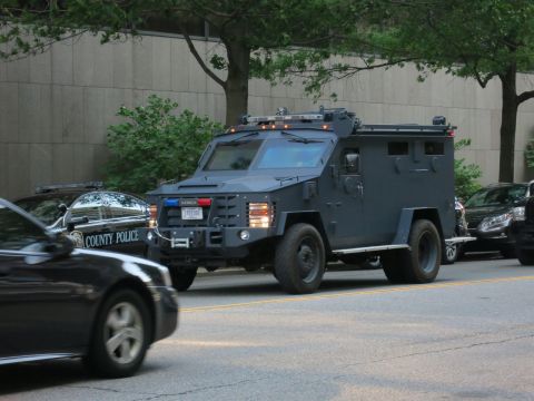 1991 Ford LTD Crown Victoria - dark gray armored tactical police vehicle drives down a street past a black County Police patrol car in the foreground.