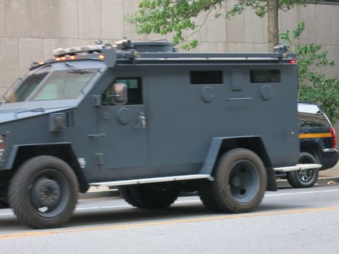 1991 Ford LTD Crown Victoria - Side profile of a dark gray armored tactical police truck with barred windows moving along a city street.