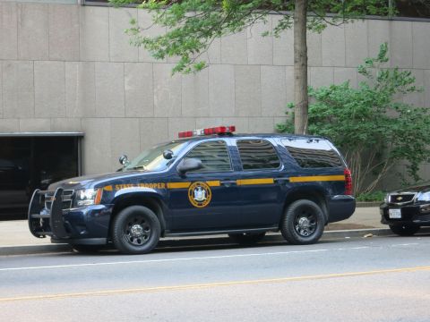 1991 Ford LTD Crown Victoria - dark blue State Trooper SUV with gold side stripe and roof lightbar parked curbside beside a stone building.