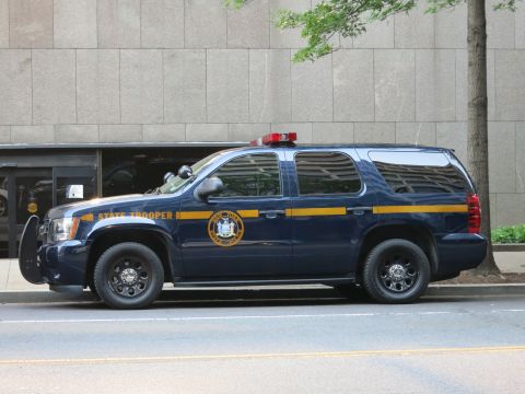 1991 Ford LTD Crown Victoria - dark blue State Trooper SUV bearing a gold stripe and seal sits parked along a downtown curb.