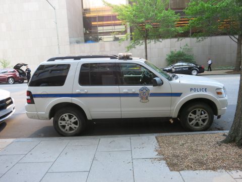1991 Ford LTD Crown Victoria - Side view of a white police SUV with blue stripe and badge parked on a city street near other patrol cars.