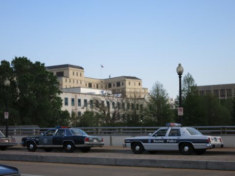 1991 Ford LTD Crown Victoria - Two vintage police sedans, one marked Boston Police, parked on a bridge with government buildings and a flag behind.