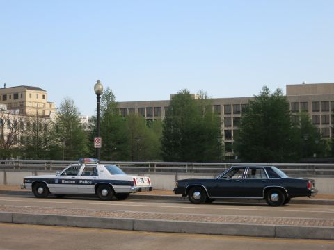 1991 Ford LTD Crown Victoria - white-and-blue Boston Police sedan beside a dark sedan on a bridge with a large building in the background.