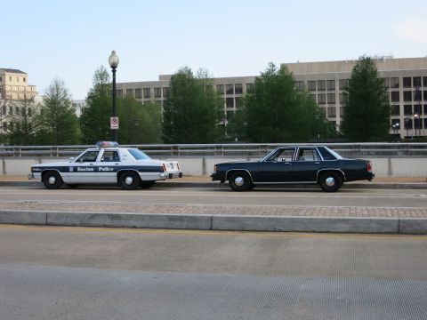 1991 Ford LTD Crown Victoria - white-and-blue Boston Police sedan next to a dark sedan on a bridge in front of a large building.