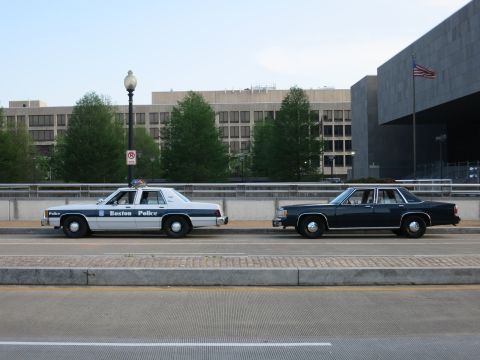1991 Ford LTD Crown Victoria - white-and-blue Boston Police sedan and a dark sedan parked on a bridge with buildings and a flag behind.