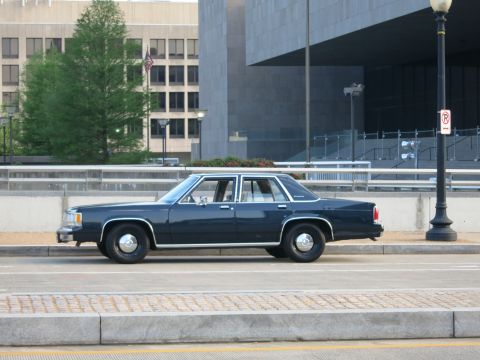 1991 Ford LTD Crown Victoria - dark blue vintage four-door sedan parked along a bridge with concrete buildings and a flag behind.