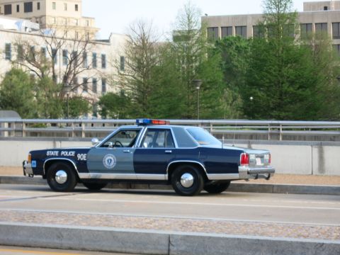 1991 Ford LTD Crown Victoria - two-tone blue State Police sedan unit 90E with agency seal parked on a bridge before city buildings.