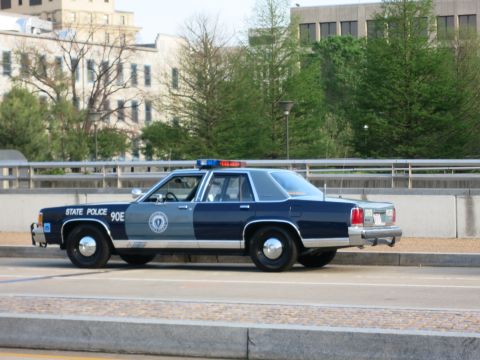 1991 Ford LTD Crown Victoria - two-tone blue State Police sedan marked 90E with an agency seal parked along a bridge near bare trees.