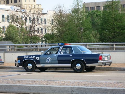 1991 Ford LTD Crown Victoria - two-tone navy and gray State Police sedan marked 90E sits on a curbed urban street with office buildings behind it.
