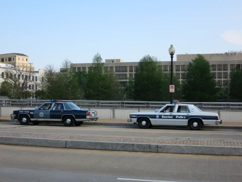 1991 Ford LTD Crown Victoria - dark State Police sedan and a white Boston Police cruiser sit nose-to-tail on an overpass with a large building behind.