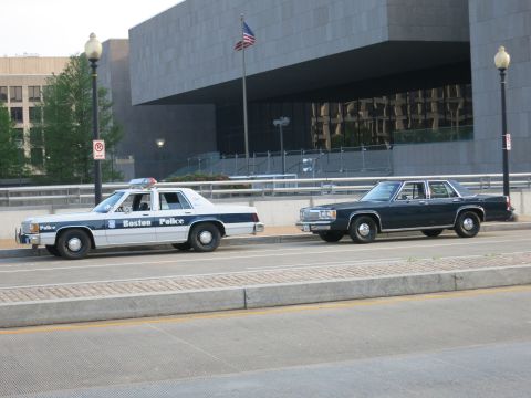 1991 Ford LTD Crown Victoria - white Boston Police cruiser and an unmarked dark sedan sit on a street fronting a stone building flying a U.S. flag.