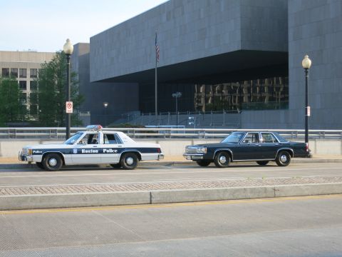 1991 Ford LTD Crown Victoria - white Boston Police cruiser parked ahead of a dark four-door sedan on a street before a stone government building.