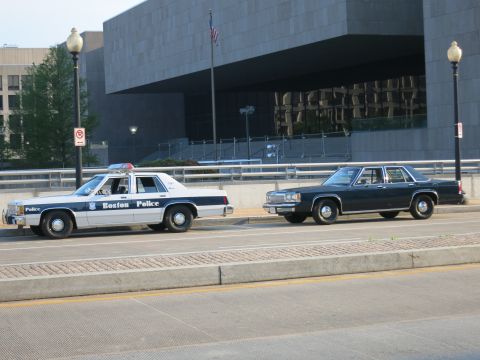 1991 Ford LTD Crown Victoria - white Boston Police cruiser and a dark sedan with a rear door ajar sit on a street before a stone building.