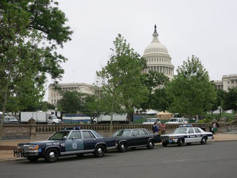 1991 Ford LTD Crown Victoria - navy State Police sedan, a dark sedan, and a Boston Police cruiser sit curbside with the U.S. Capitol dome rising behind trees.