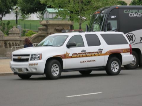 1991 Ford LTD Crown Victoria - white Chevrolet Suburban marked Sheriff Alexandria sits in front of a Campus Coach motorcoach near a stone wall.