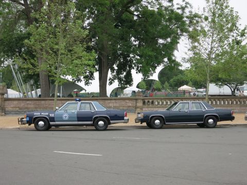 1991 Ford LTD Crown Victoria - navy State Police sedan marked 90E sits ahead of a dark four-door sedan on a tree-lined street near tents.