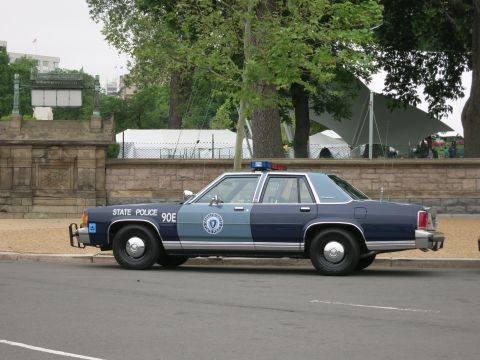 1991 Ford LTD Crown Victoria - two-tone navy and gray State Police sedan marked 90E is parked in profile beside a stone wall and white event tent.