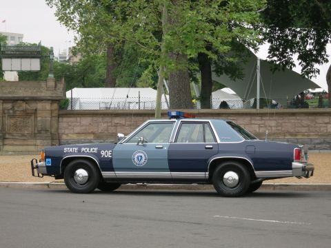 1991 Ford LTD Crown Victoria - two-tone State Police sedan marked 90E with a push bar sits in side profile near a stone wall and tents.