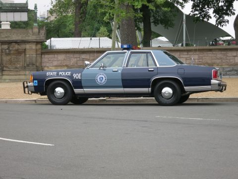 1991 Ford LTD Crown Victoria - two-tone navy and gray State Police sedan marked 90E is parked in profile along a curb by a stone wall.