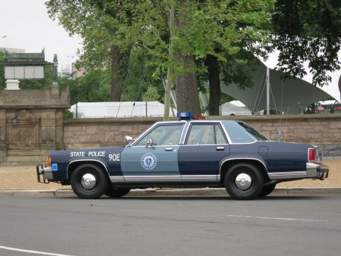 1991 Ford LTD Crown Victoria - two-tone State Police sedan marked 90E sits in side profile beside a stone wall with event tents behind.