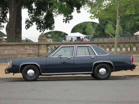 1991 Ford LTD Crown Victoria - dark blue full-size four-door sedan with chrome trim and hubcaps sits in profile along a curb near event tents.