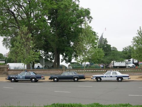 1991 Ford LTD Crown Victoria - navy sedan, a dark sedan, and a white Boston Police cruiser sit in a row on a street with trees and tents behind.