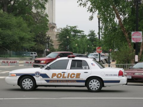 1991 Ford LTD Crown Victoria - white United States Capitol Police K-9 cruiser drives a city street past a red pickup, stop barriers, and trees.
