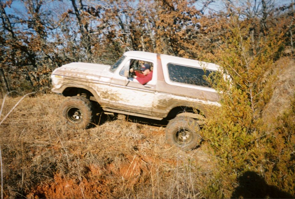 Lifted white late-70s Ford Bronco streaked with mud climbing a red-dirt trail through brush