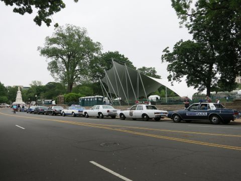 1991 Ford LTD Crown Victoria - long line of older police cars and sedans parked along a roadside in front of a white tent canopy and trees.
