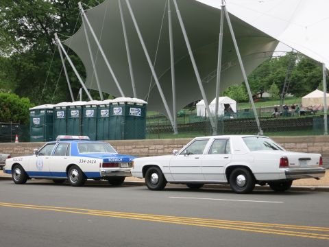 1991 Ford LTD Crown Victoria - blue and white Norwich police sedan parked next to a plain white sedan in front of green portable toilets and a canopy.