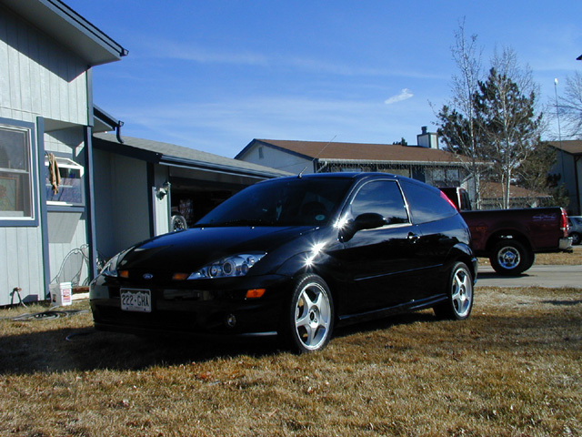 Black 3-door Ford Focus hatchback parked in front of a house, with a red pickup truck in the background