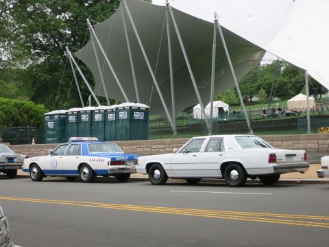 1991 Ford LTD Crown Victoria - blue and white police car marked 334-2424 parked beside a white sedan in front of a sweeping white canopy.