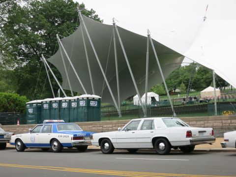 1991 Ford LTD Crown Victoria - blue and white police sedan and a white four-door sedan parked at a curb beneath a large white tent canopy.