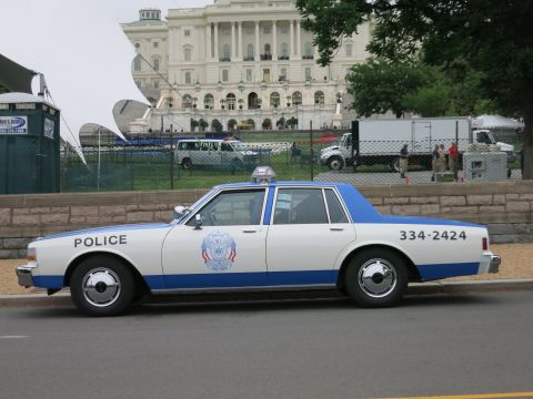 1991 Ford LTD Crown Victoria - Side profile of a blue and white police car marked 334-2424 parked along a fence with the Capitol building behind.