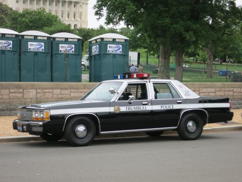 1991 Ford LTD Crown Victoria - black and white Trumbull Police car numbered 13 parked at a curb in front of green portable toilets and a fenced lawn.