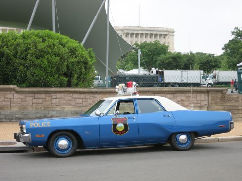 1991 Ford LTD Crown Victoria - blue and white vintage Police sedan with roof light parked beside a stone planter wall with trees behind.