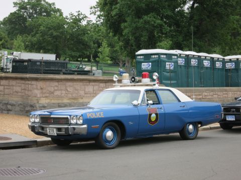 1991 Ford LTD Crown Victoria - blue and white vintage Police sedan numbered 218 with twin roof lights parked beside a stone wall and portable toilets.