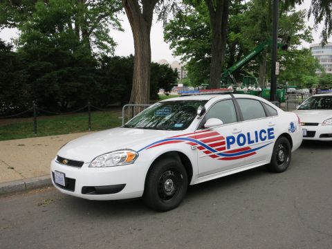 1991 Ford LTD Crown Victoria - white Chevrolet Impala police sedan with red and blue swoosh striping and POLICE lettering parked on a tree-lined street.