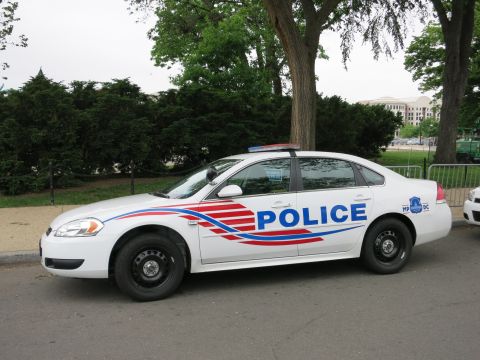 1991 Ford LTD Crown Victoria - Driver-side view of a white Chevrolet Impala police sedan with red and blue striping and POLICE lettering parked by trees.