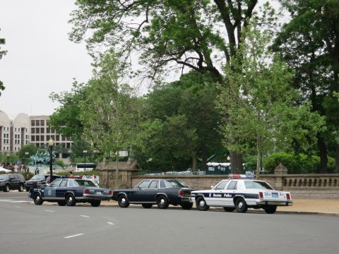 1991 Ford LTD Crown Victoria - row of classic full-size police sedans, including a Boston Police car, parked along a street in front of a large building.